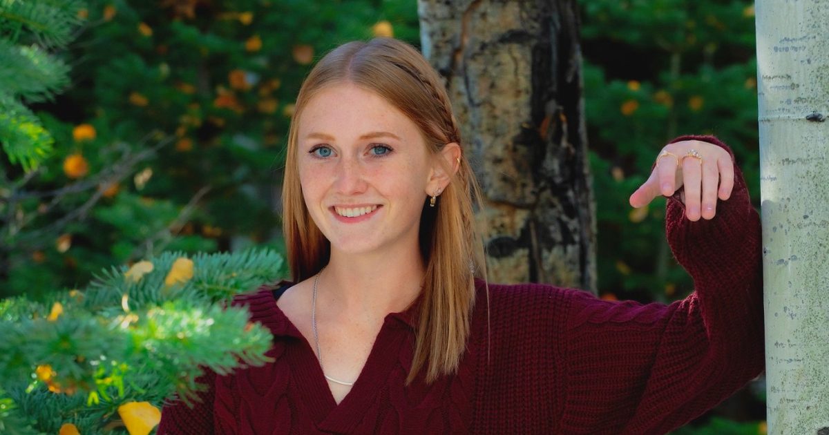 A young woman with red hair, wearing a maroon sweater, smiles while leaning against a tree in a forest setting.