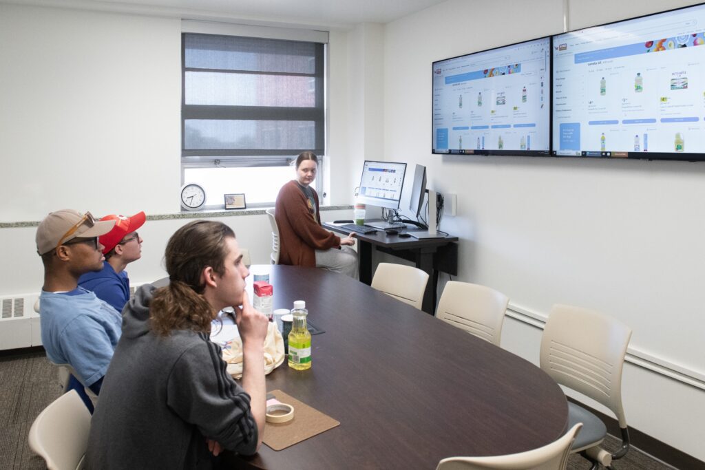 A classroom setting with several people seated around a table, watching a presentation on two large screens. The screens display an online shopping interface. A presenter sits at a computer desk, guiding the session.