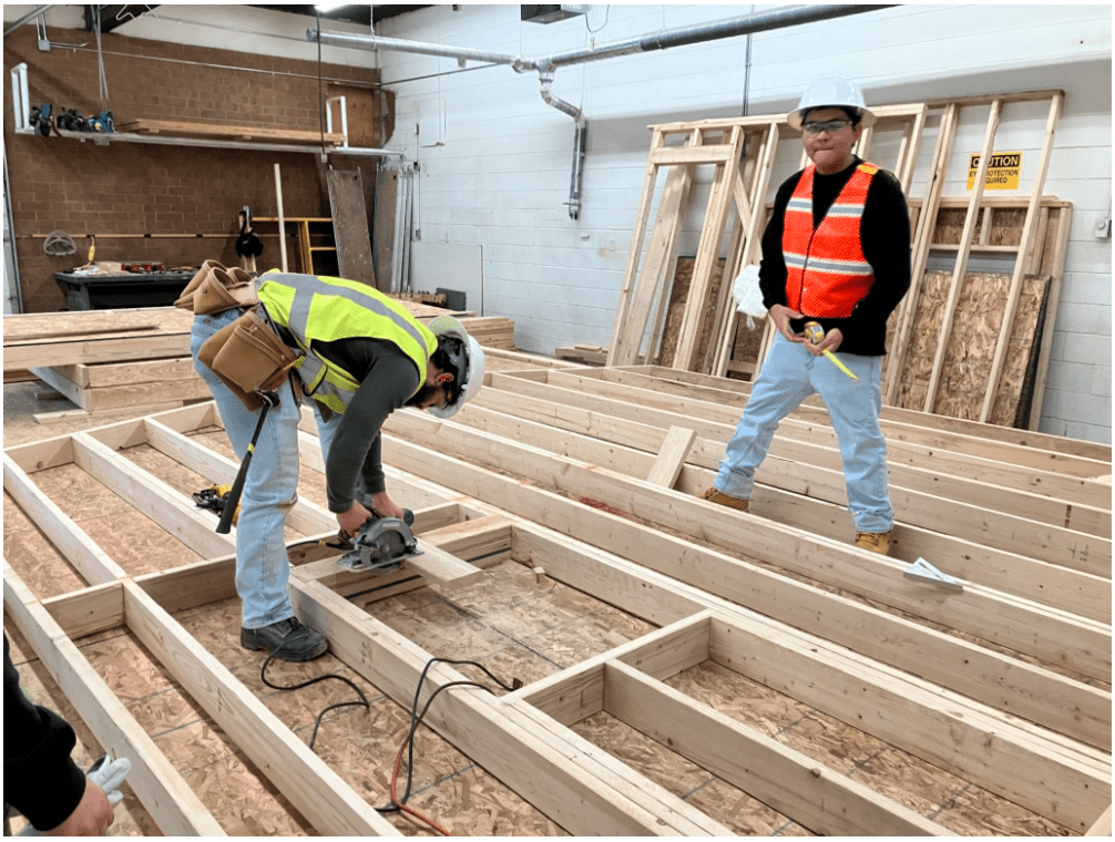 Two construction students are working on framing a wooden structure indoors. One worker is using a circular saw to cut a piece of wood, while the other stands nearby holding a tape measure. Both are wearing safety helmets and high-visibility vests.