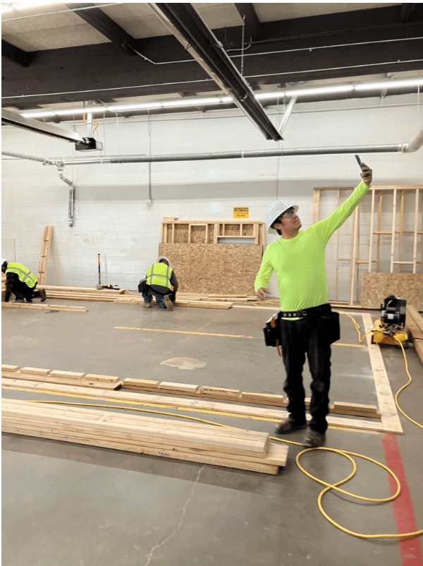 A construction worker wearing a bright green shirt and safety helmet is taking a selfie inside a large indoor workspace. Other workers in the background are assembling wooden frames.