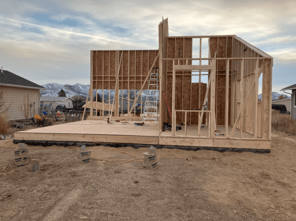 An outdoor construction site showing the framework of a building under construction. The structure has partial walls in place, and the background features mountains under a cloudy sky.