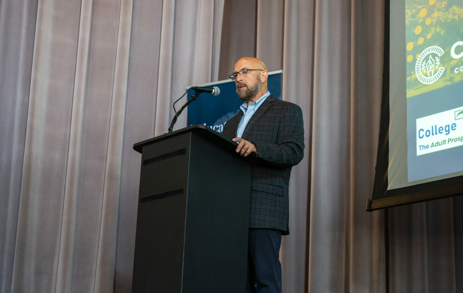 A man stands at a podium speaking into a microphone. Behind him, a banner partially visible reads, 'Education that meets students where they are.' The background also includes a projection screen with logos and text related to the event.