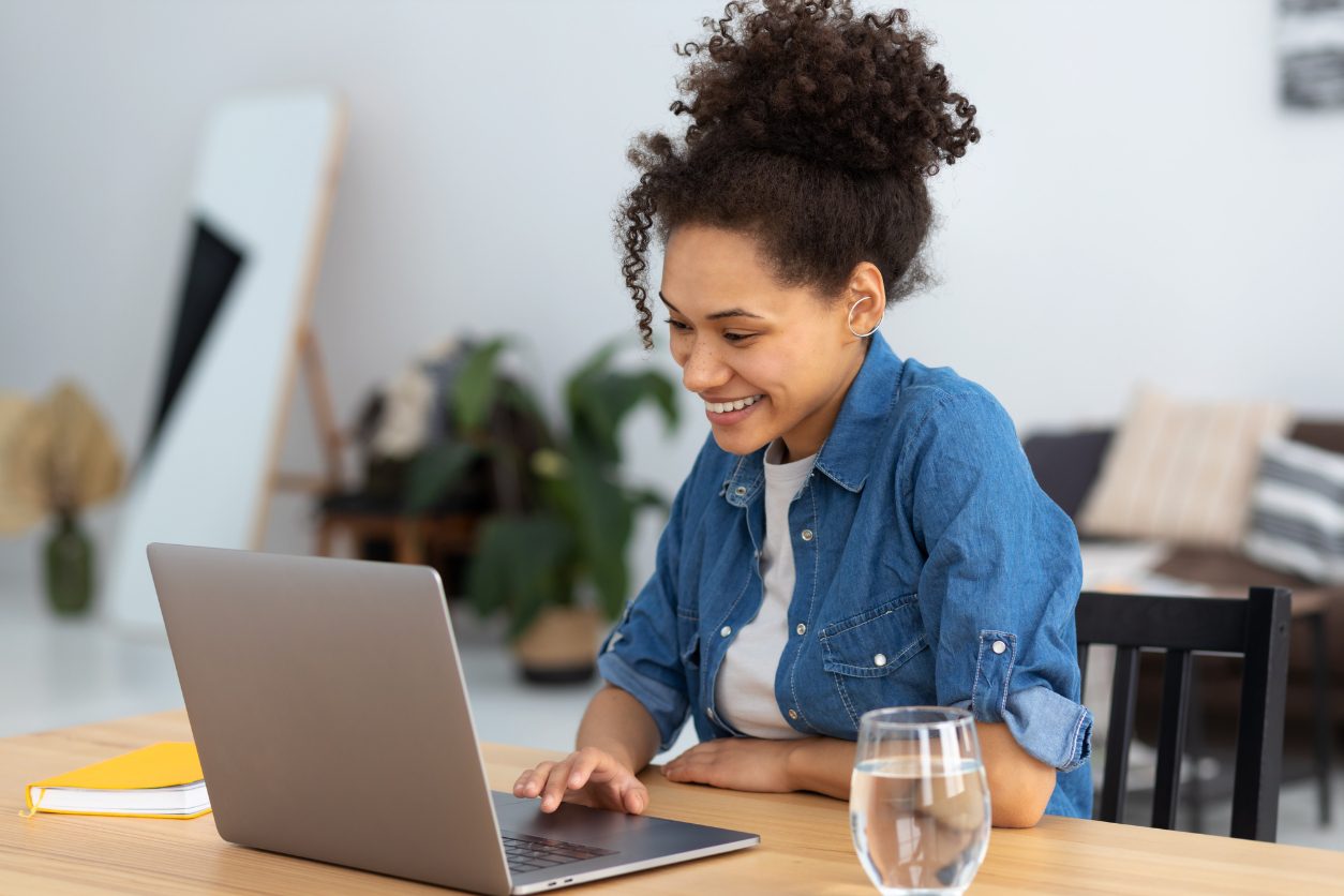 Smiling woman working on a laptop at a table with a glass of water and a yellow notebook in a cozy, well-decorated room.