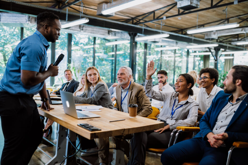 A diverse group of professionals engaged in a business meeting in a bright, modern office. One woman is raising her hand, possibly to ask a question, while the presenter speaks into a microphone.