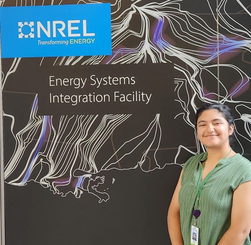 Community College of Denver student Emily Serrano stands in front of a sign that says "NREL: Energy Systems Integration Facility." Emily is a young Latina woman wearing a textured sage green top.