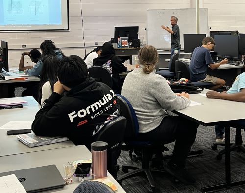 Community College of Denver faculty member Jason Burke stands at the front of the classroom leading a math class of ten students. Jason is wearing a gray polo shirt and jeans. The classroom is white with a bank of black computers.