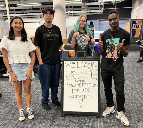 Four CCD students stand in front of a white board that says, "Welcome Summer Engineering Bridge Students!" Left to right, the students are a young Latina woman wearing a white top and jean shorts; a young Latino man wearing a black t-shirt and black jeans; a white, blond woman wearing a tie-dye shirt; and a young Black man wearing a black t-shirt and black jeans.