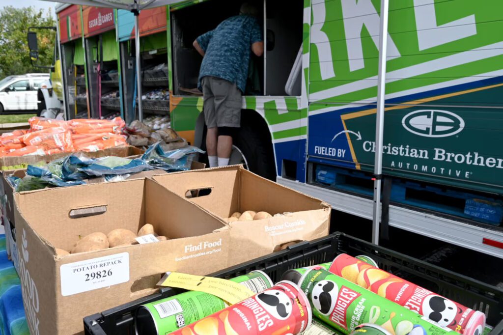 A community food distribution event where boxes of fresh produce, including potatoes and carrots, are displayed on a table. In the background, a person in a blue shirt and gray shorts is loading or unloading supplies from a large, brightly colored truck labeled with "Fueled by Christian Brothers Automotive."