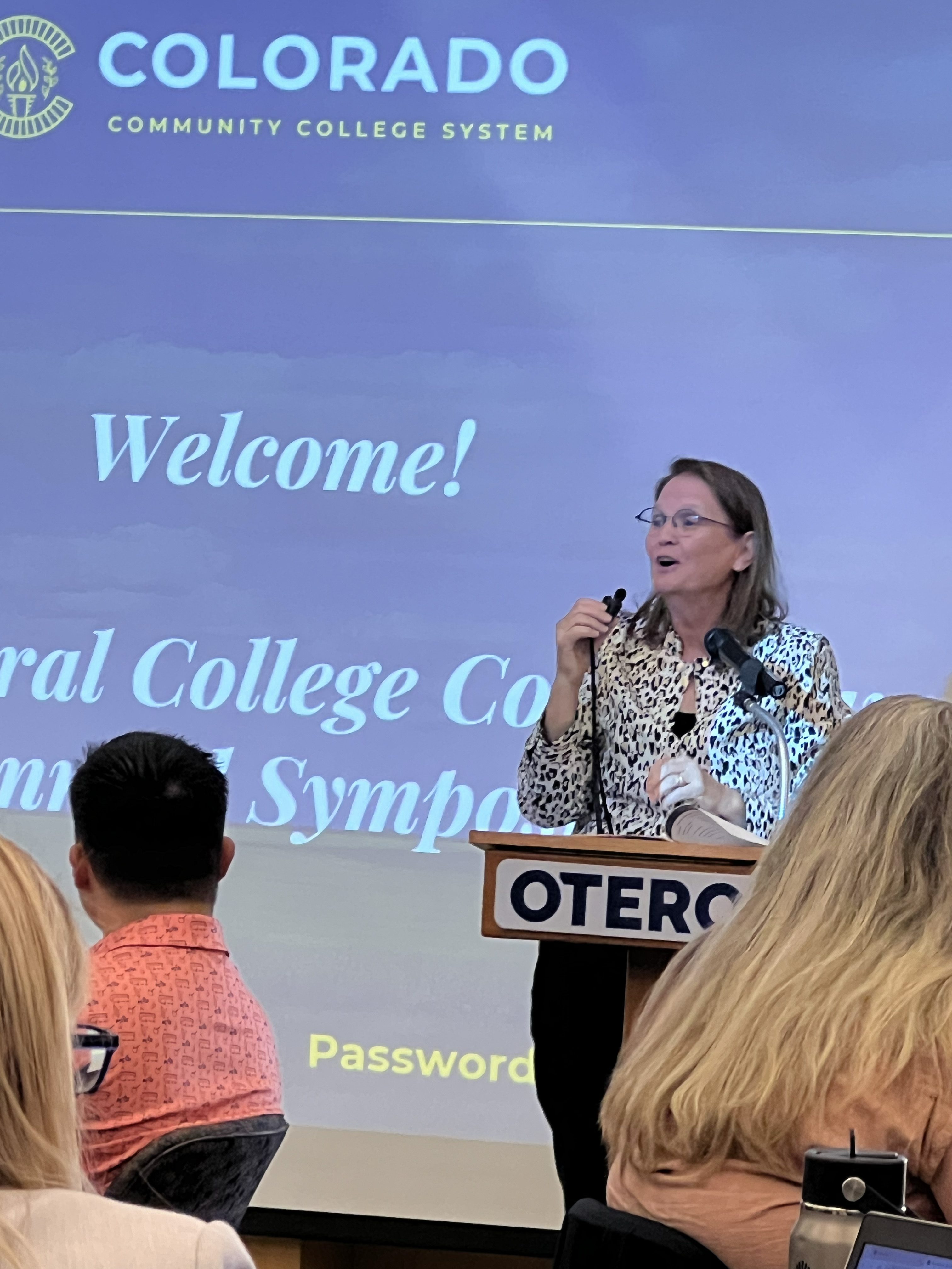 Diane Duffy stands at a podium labeled "OTERO," addressing the audience in front of a large screen that reads "Welcome! Rural College Conference Annual Symposium" with the Colorado Community College System logo.