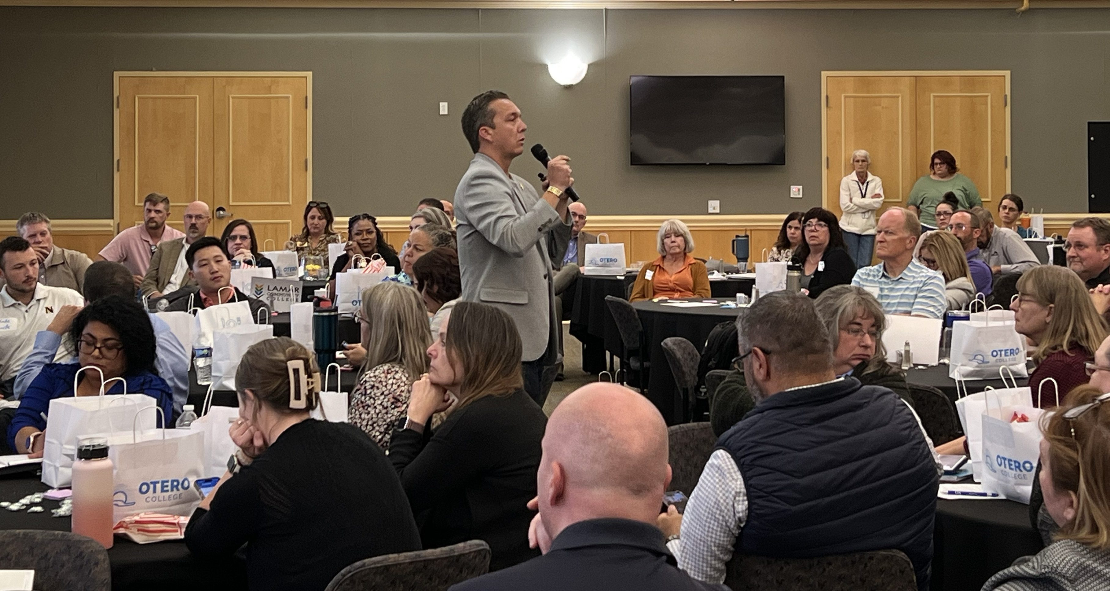 Landon Mascareñaz stands in the middle of a crowded conference room, holding a microphone and engaging with attendees seated at round tables. The audience members appear attentive, with conference materials and tote bags on the tables.