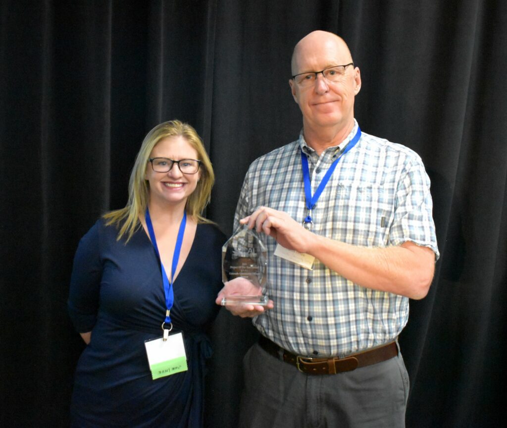 A man and a woman stand together on a stage, smiling at the camera. The man is holding a clear glass award, while both are wearing lanyards and name badges.