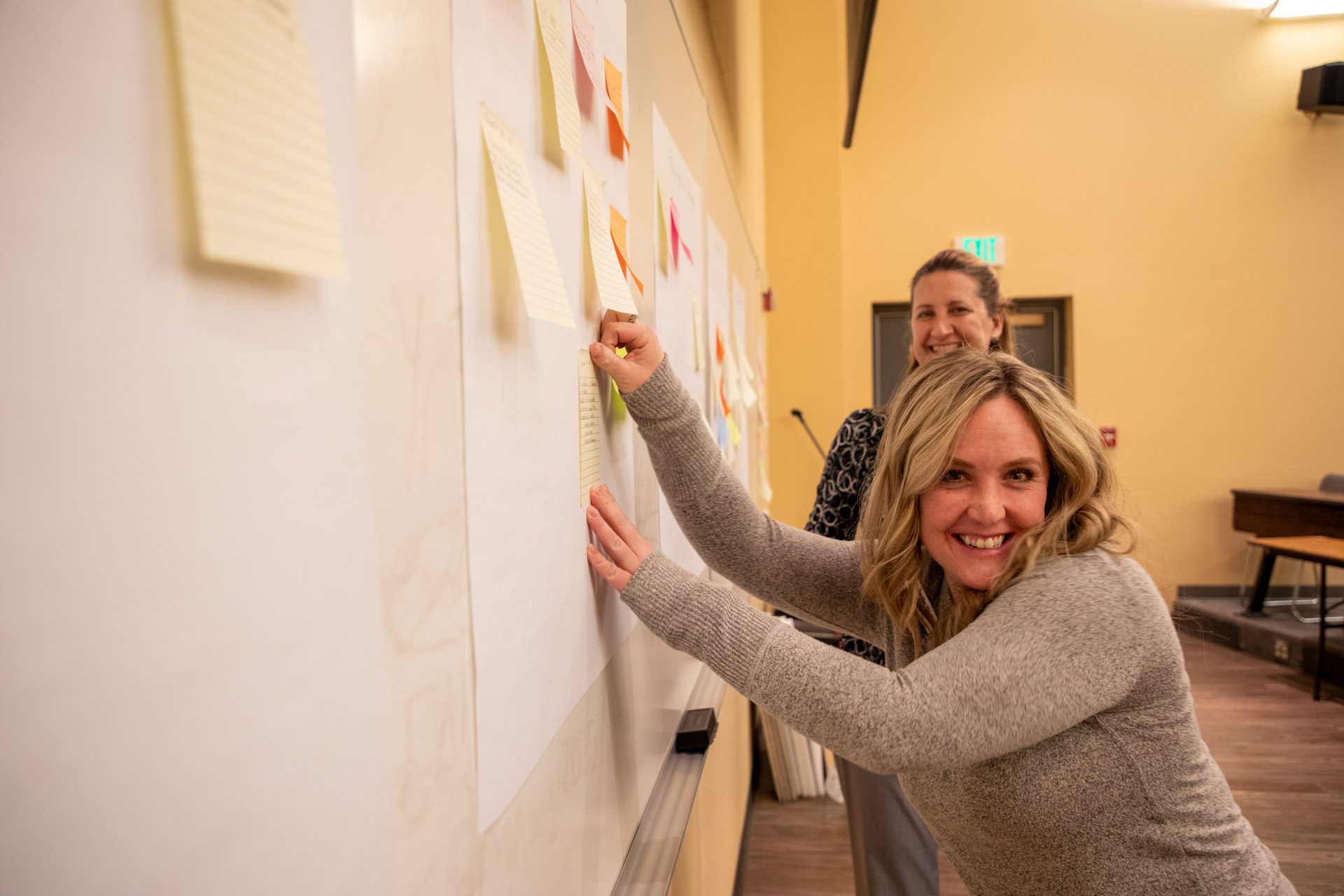 two women in a classroom smiling while participating in a workshop with sticky notes and pads on the wall